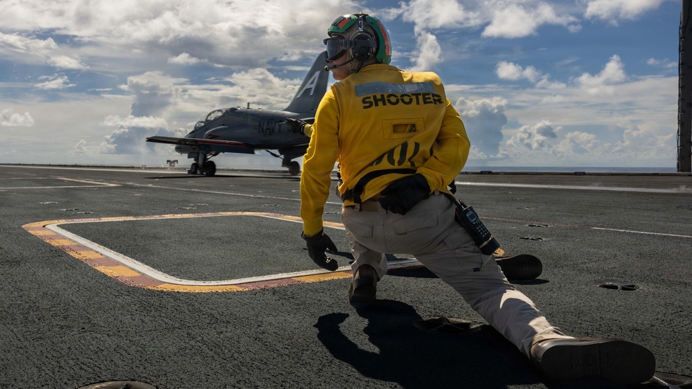 A U.S. Navy 'Shooter' (catapult officer) in a yellow jersey is crouched in the characteristic launch-signal pose on an aircraft carrier flight deck. A Navy training aircraft, identifiable as a T-45 Goshawk marked 'NAVY,' is positioned on the catapult track ahead of them. The scene takes place at sea under a partly cloudy sky.