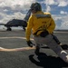 A U.S. Navy 'Shooter' (catapult officer) in a yellow jersey is crouched in the characteristic launch-signal pose on an aircraft carrier flight deck. A Navy training aircraft, identifiable as a T-45 Goshawk marked 'NAVY,' is positioned on the catapult track ahead of them. The scene takes place at sea under a partly cloudy sky.