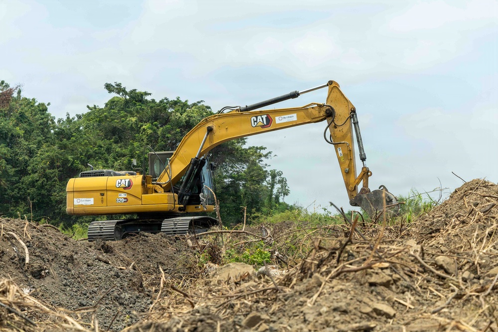 A yellow Caterpillar (CAT) 320 excavator is actively working on a land clearing or earthmoving site. The machine is positioned on disturbed soil surrounded by debris, uprooted vegetation, and mounds of earth. Dense green forest vegetation is visible in the background, suggesting this may be a deforestation or site preparation operation.