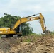 A yellow Caterpillar (CAT) 320 excavator is actively working on a land clearing or earthmoving site. The machine is positioned on disturbed soil surrounded by debris, uprooted vegetation, and mounds of earth. Dense green forest vegetation is visible in the background, suggesting this may be a deforestation or site preparation operation.