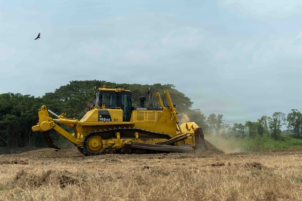 A large yellow Komatsu D155AX bulldozer is actively grading or clearing land in an open field. The machine is pushing soil and raising dust as it operates, with a treeline visible in the background. A bird is visible in flight in the upper left portion of the sky.
