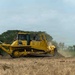 A large yellow Komatsu D155AX bulldozer is actively grading or clearing land in an open field. The machine is pushing soil and raising dust as it operates, with a treeline visible in the background. A bird is visible in flight in the upper left portion of the sky.