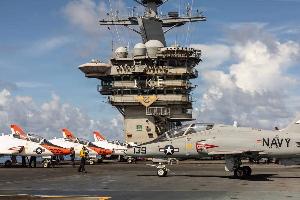 The image shows the flight deck of USS Dwight D. Eisenhower (CVN-69), identifiable by the 'IKE' marking on the island superstructure. Multiple T-45 Goshawk trainer aircraft in white and orange Navy livery are parked on the deck, with visible tail numbers including 235, 162, and 139. Deck crew in yellow jerseys are visible conducting flight deck operations.