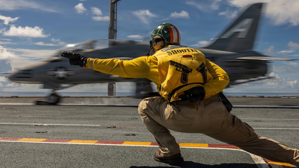 A U.S. Navy 'Shooter' (catapult officer) in a yellow jersey and green helmet crouches in the classic launch signal pose on an aircraft carrier flight deck. A military jet aircraft, bearing U.S. roundel markings, is captured mid-launch in a motion-blurred streak across the frame. The image conveys the high-energy, dynamic environment of carrier flight operations.