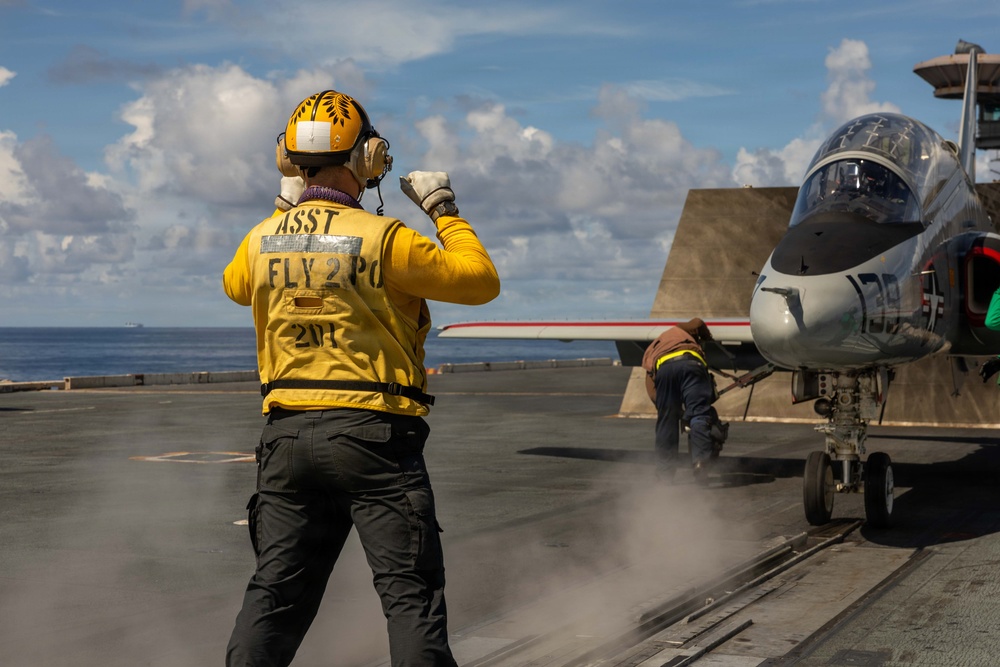 A flight deck crew member wearing a yellow jersey labeled 'ASST FLY 2 PO 201' signals on the flight deck of a naval aircraft carrier. A military jet aircraft, likely an F/A-18 Hornet or similar carrier-based fighter, is positioned on the catapult with steam visible beneath it, indicating launch preparation. A second crew member is visible near the aircraft's landing gear.
