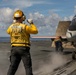 A flight deck crew member wearing a yellow jersey labeled 'ASST FLY 2 PO 201' signals on the flight deck of a naval aircraft carrier. A military jet aircraft, likely an F/A-18 Hornet or similar carrier-based fighter, is positioned on the catapult with steam visible beneath it, indicating launch preparation. A second crew member is visible near the aircraft's landing gear.