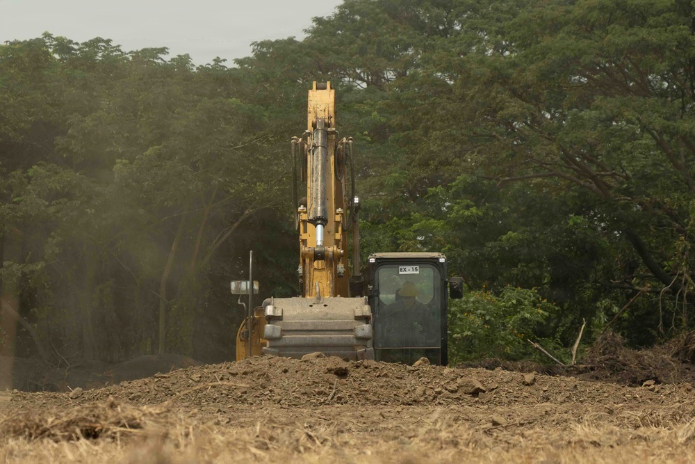 A yellow hydraulic excavator, marked 'EX-15', is actively engaged in earthmoving or land-clearing operations on a dusty, barren plot of land. The machine is partially obscured by a mound of disturbed soil in the foreground. Dense green trees form the background, suggesting the clearing is occurring at the edge of a forested or vegetated area.