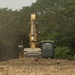 A yellow hydraulic excavator, marked 'EX-15', is actively engaged in earthmoving or land-clearing operations on a dusty, barren plot of land. The machine is partially obscured by a mound of disturbed soil in the foreground. Dense green trees form the background, suggesting the clearing is occurring at the edge of a forested or vegetated area.