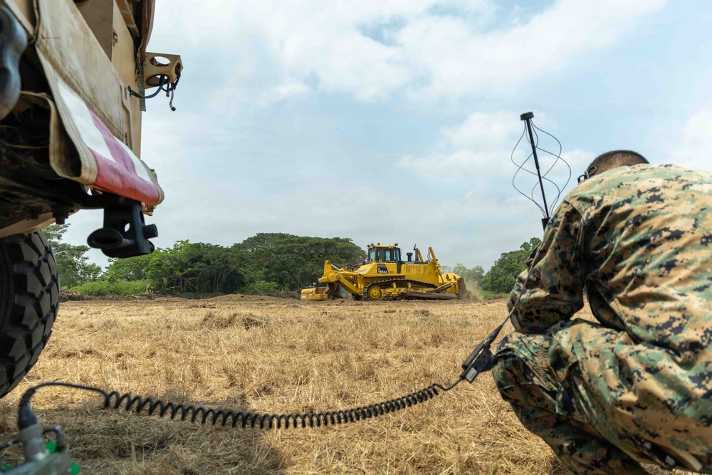 A military service member in MARPAT camouflage uniform crouches beside a tactical vehicle while observing a yellow bulldozer (appears to be a Komatsu D65 or similar model) operating in an open dry field. The scene appears to depict a military engineering or construction operation, likely airfield or road preparation. The service member is holding a radio handset connected to a coiled cable, suggesting communications coordination.