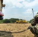 A military service member in MARPAT camouflage uniform crouches beside a tactical vehicle while observing a yellow bulldozer (appears to be a Komatsu D65 or similar model) operating in an open dry field. The scene appears to depict a military engineering or construction operation, likely airfield or road preparation. The service member is holding a radio handset connected to a coiled cable, suggesting communications coordination.