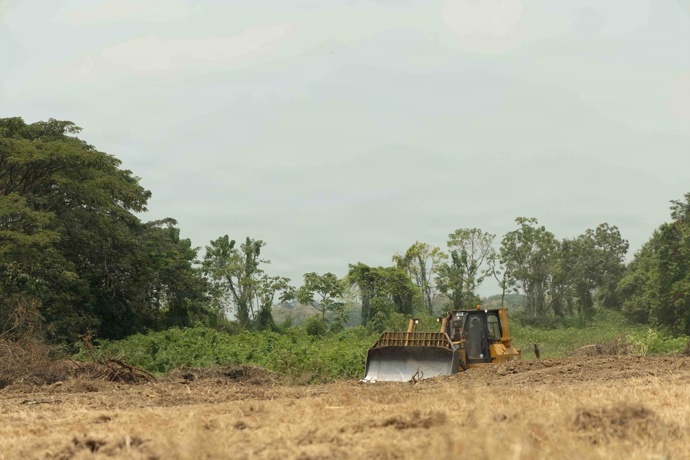 A yellow Caterpillar bulldozer is actively clearing land in what appears to be a tropical or subtropical region. The foreground shows already-cleared, dry earth while the background retains dense green vegetation and trees. This image depicts active land clearing or deforestation activity.