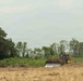 A yellow Caterpillar bulldozer is actively clearing land in what appears to be a tropical or subtropical region. The foreground shows already-cleared, dry earth while the background retains dense green vegetation and trees. This image depicts active land clearing or deforestation activity.