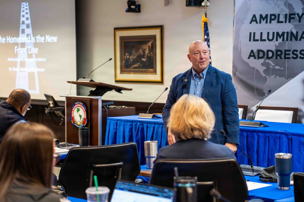 A bald, older male speaker in a blue blazer and checkered shirt addresses an audience at a formal conference or briefing session. The presentation slide behind him reads 'The Homeland Is the New Front Line,' suggesting a national security or defense-related topic. The setting includes a blue-draped head table, a podium with an organizational seal, and a banner partially reading 'AMPLIFY, ILLUMINA[TE], ADDRESS.'