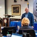 A bald, older male speaker in a blue blazer and checkered shirt addresses an audience at a formal conference or briefing session. The presentation slide behind him reads 'The Homeland Is the New Front Line,' suggesting a national security or defense-related topic. The setting includes a blue-draped head table, a podium with an organizational seal, and a banner partially reading 'AMPLIFY, ILLUMINA[TE], ADDRESS.'