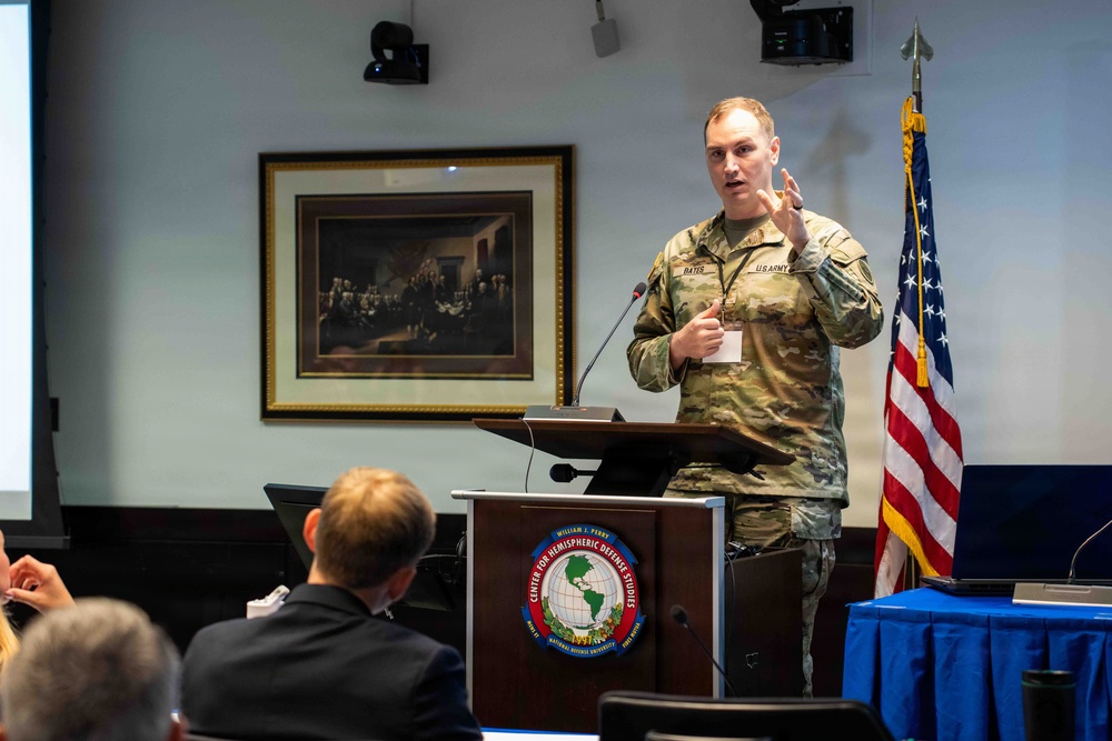 A U.S. Army soldier is presenting at a podium bearing the seal of the William J. Perry Center for Hemispheric Defense Studies at the National Defense University. The speaker, whose name tag reads 'BATES,' is in military camouflage uniform and is gesturing while addressing an audience. An American flag and a framed painting resembling the signing of the Declaration of Independence are visible in the background.