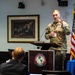 A U.S. Army soldier is presenting at a podium bearing the seal of the William J. Perry Center for Hemispheric Defense Studies at the National Defense University. The speaker, whose name tag reads 'BATES,' is in military camouflage uniform and is gesturing while addressing an audience. An American flag and a framed painting resembling the signing of the Declaration of Independence are visible in the background.
