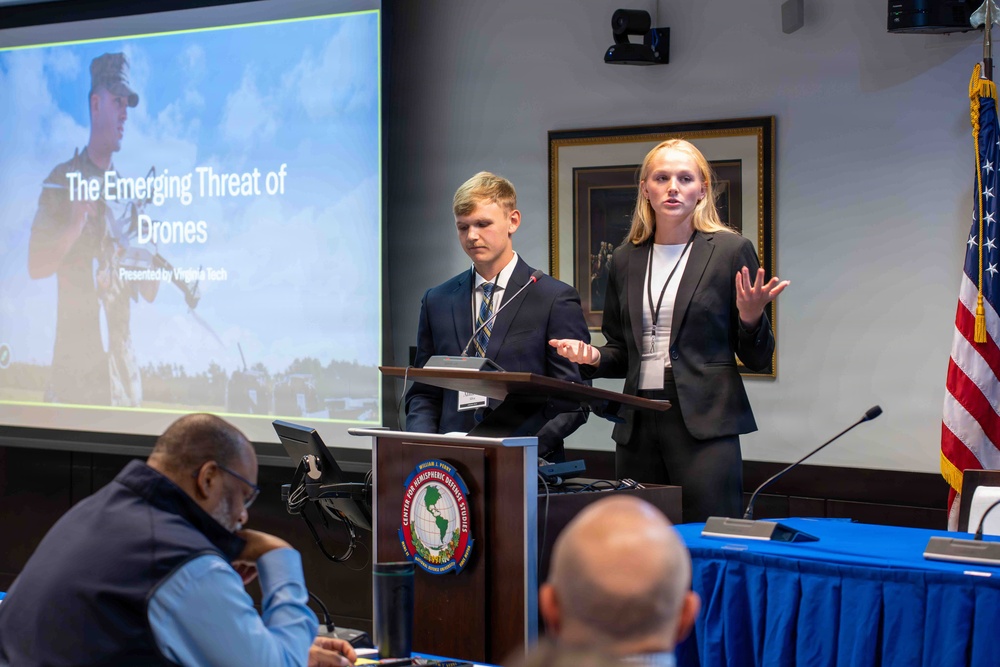 Two young presenters, a male and a female, are delivering a presentation titled 'The Emerging Threat of Drones' on behalf of Virginia Tech at what appears to be a formal defense or security conference. They are standing at a podium bearing the seal of the Center for Hemispheric Defense Studies, with an audience seated in front of them. The setting is a professional conference room with a U.S. flag and projection screen visible.