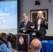 Two young presenters, a male and a female, are delivering a presentation titled 'The Emerging Threat of Drones' on behalf of Virginia Tech at what appears to be a formal defense or security conference. They are standing at a podium bearing the seal of the Center for Hemispheric Defense Studies, with an audience seated in front of them. The setting is a professional conference room with a U.S. flag and projection screen visible.