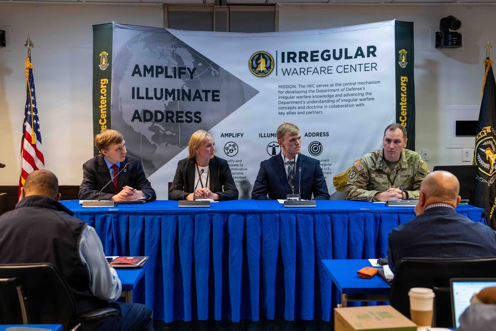 A panel discussion is taking place at an Irregular Warfare Center (IWC) event, with four panelists seated at a blue-draped table in front of a large branded backdrop. The panel includes three civilians (two men and one woman) and one U.S. Army soldier in military uniform. Audience members are visible from behind in the foreground, with some using laptops and taking notes.