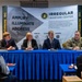 A panel discussion is taking place at an Irregular Warfare Center (IWC) event, with four panelists seated at a blue-draped table in front of a large branded backdrop. The panel includes three civilians (two men and one woman) and one U.S. Army soldier in military uniform. Audience members are visible from behind in the foreground, with some using laptops and taking notes.