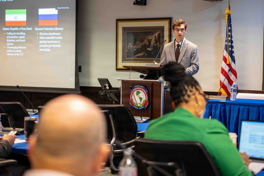 A young man in a gray suit is presenting at a podium bearing the seal of the Center for Hemispheric Defense Studies (William J. Perry Center) to an audience seated at conference tables. The presentation slide visible behind him displays flags and bullet points about Iran and Russia, covering topics such as election interference and state-controlled media. The setting appears to be a formal academic or government conference room with a U.S. flag and a historical painting on the wall.