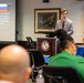 A young man in a gray suit is presenting at a podium bearing the seal of the Center for Hemispheric Defense Studies (William J. Perry Center) to an audience seated at conference tables. The presentation slide visible behind him displays flags and bullet points about Iran and Russia, covering topics such as election interference and state-controlled media. The setting appears to be a formal academic or government conference room with a U.S. flag and a historical painting on the wall.