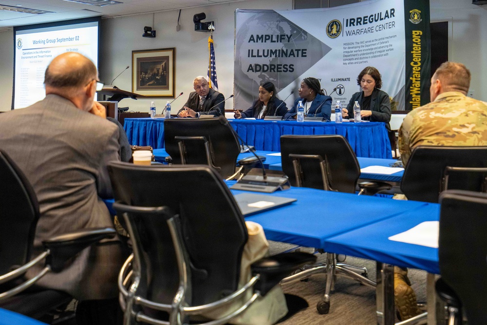 A formal working group session is being conducted under the banner of the Irregular Warfare Center, with a panel of four individuals seated at a blue-draped table facing an audience. The event appears to be a structured briefing or discussion session, with a presentation slide visible on a screen to the left labeled 'Working Group September 02'. Attendees including civilians and at least one military personnel in camouflage uniform are seated at blue-covered tables facing the panel.