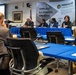 A formal working group session is being conducted under the banner of the Irregular Warfare Center, with a panel of four individuals seated at a blue-draped table facing an audience. The event appears to be a structured briefing or discussion session, with a presentation slide visible on a screen to the left labeled 'Working Group September 02'. Attendees including civilians and at least one military personnel in camouflage uniform are seated at blue-covered tables facing the panel.