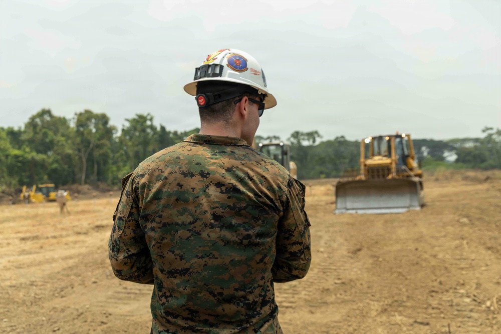 A military service member wearing digital camouflage (MARPAT pattern) and a white hard hat with a unit insignia patch is observing construction equipment operations on a cleared dirt field. At least two yellow bulldozers are visible operating in the background, with dense tropical vegetation lining the perimeter. This appears to be a military engineering or construction operation, likely a site clearing or grading project.