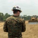 A military service member wearing digital camouflage (MARPAT pattern) and a white hard hat with a unit insignia patch is observing construction equipment operations on a cleared dirt field. At least two yellow bulldozers are visible operating in the background, with dense tropical vegetation lining the perimeter. This appears to be a military engineering or construction operation, likely a site clearing or grading project.