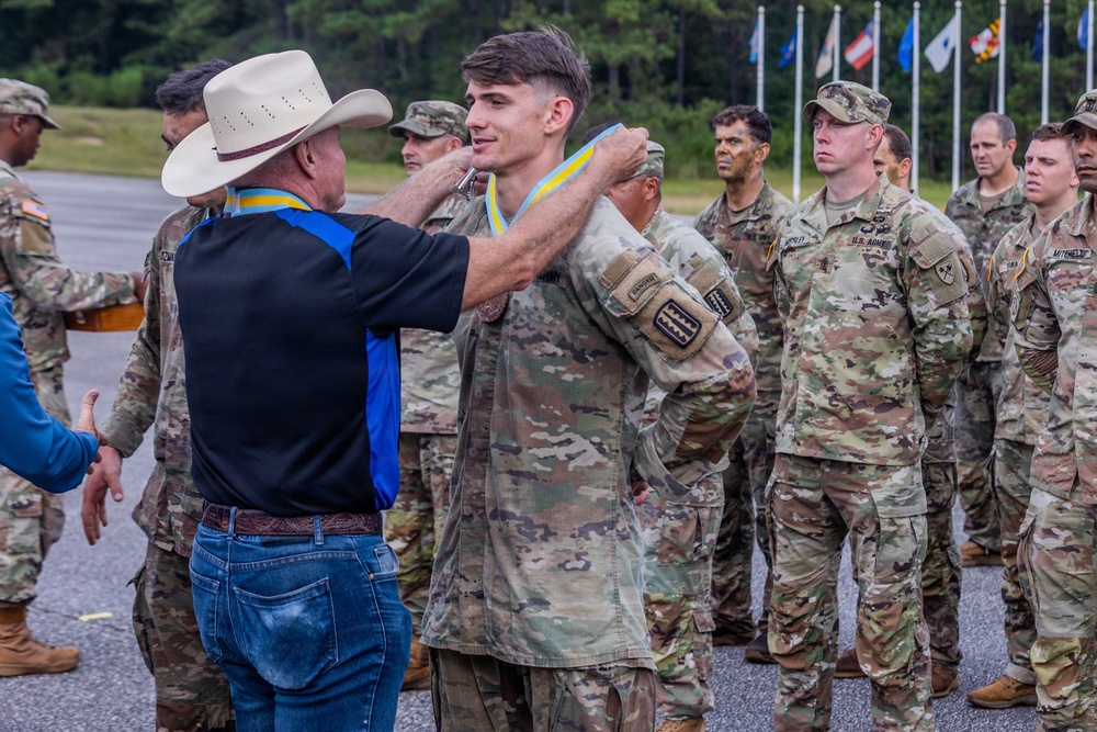 A civilian man wearing a white cowboy hat, black shirt, and jeans is placing a blue and yellow medal/ribbon around the neck of a U.S. Army soldier in OCP (Operational Camouflage Pattern) uniform. The soldier appears to be receiving an award or recognition during a formal military ceremony. Multiple other soldiers in uniform are standing in formation in the background.
