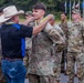 A civilian man wearing a white cowboy hat, black shirt, and jeans is placing a blue and yellow medal/ribbon around the neck of a U.S. Army soldier in OCP (Operational Camouflage Pattern) uniform. The soldier appears to be receiving an award or recognition during a formal military ceremony. Multiple other soldiers in uniform are standing in formation in the background.