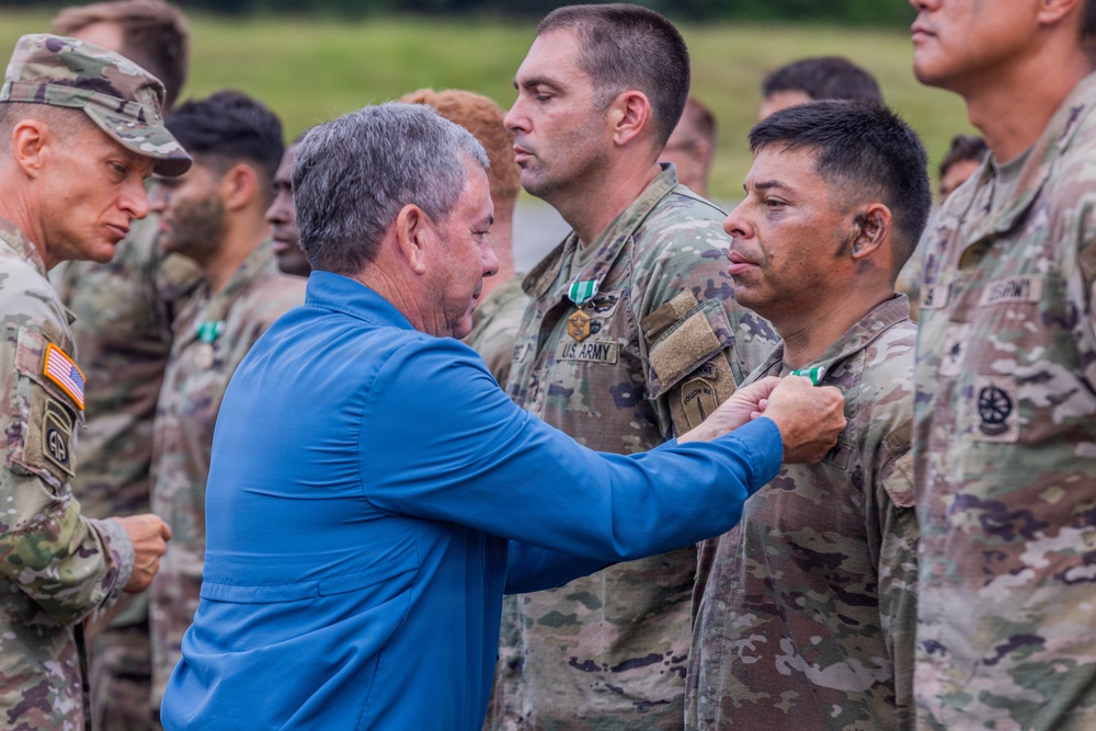 A civilian in a blue shirt is pinning a medal or award onto a U.S. Army soldier during what appears to be a military awards ceremony conducted outdoors. Multiple soldiers in OCP (Operational Camouflage Pattern) uniforms are standing in formation in the background. At least one other soldier in the background is already wearing a green medal, suggesting this is an active decoration event.