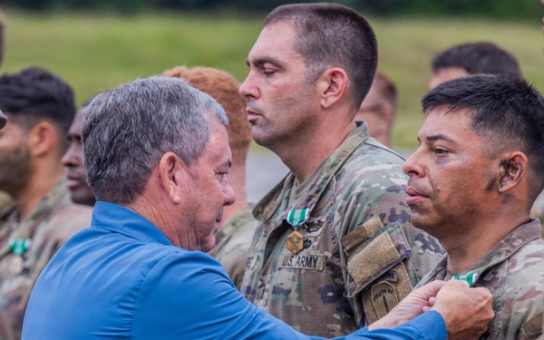 A civilian in a blue shirt is pinning a medal or award onto a U.S. Army soldier during what appears to be a military awards ceremony conducted outdoors. Multiple soldiers in OCP (Operational Camouflage Pattern) uniforms are standing in formation in the background. At least one other soldier in the background is already wearing a green medal, suggesting this is an active decoration event.