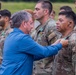 A civilian in a blue shirt is pinning a medal or award onto a U.S. Army soldier during what appears to be a military awards ceremony conducted outdoors. Multiple soldiers in OCP (Operational Camouflage Pattern) uniforms are standing in formation in the background. At least one other soldier in the background is already wearing a green medal, suggesting this is an active decoration event.