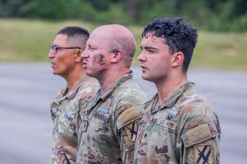 Three U.S. Army soldiers in Operational Camouflage Pattern (OCP) uniforms are standing in a line, facing the same direction in what appears to be a formation or ceremony. The middle soldier has visible face camouflage paint, suggesting recent field training or exercise activity. All three wear 'U.S. ARMY' chest tape and unit patches on their shoulders.