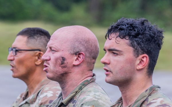 Three U.S. Army soldiers in Operational Camouflage Pattern (OCP) uniforms are standing in a line, facing the same direction in what appears to be a formation or ceremony. The middle soldier has visible face camouflage paint, suggesting recent field training or exercise activity. All three wear 'U.S. ARMY' chest tape and unit patches on their shoulders.