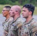 Three U.S. Army soldiers in Operational Camouflage Pattern (OCP) uniforms are standing in a line, facing the same direction in what appears to be a formation or ceremony. The middle soldier has visible face camouflage paint, suggesting recent field training or exercise activity. All three wear 'U.S. ARMY' chest tape and unit patches on their shoulders.