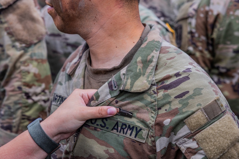 A close-up photograph showing a hand pinning or attaching a small item onto the chest area of a U.S. Army soldier's OCP (Operational Camouflage Pattern) uniform. The name tape 'U.S. ARMY' is clearly visible on the uniform. The scene appears to be a military ceremony or formal recognition event, with additional soldiers visible in the background.