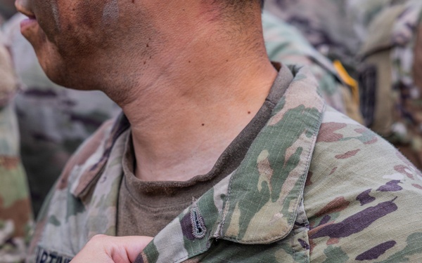 A close-up photograph showing a hand pinning or attaching a small item onto the chest area of a U.S. Army soldier's OCP (Operational Camouflage Pattern) uniform. The name tape 'U.S. ARMY' is clearly visible on the uniform. The scene appears to be a military ceremony or formal recognition event, with additional soldiers visible in the background.