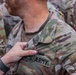 A close-up photograph showing a hand pinning or attaching a small item onto the chest area of a U.S. Army soldier's OCP (Operational Camouflage Pattern) uniform. The name tape 'U.S. ARMY' is clearly visible on the uniform. The scene appears to be a military ceremony or formal recognition event, with additional soldiers visible in the background.
