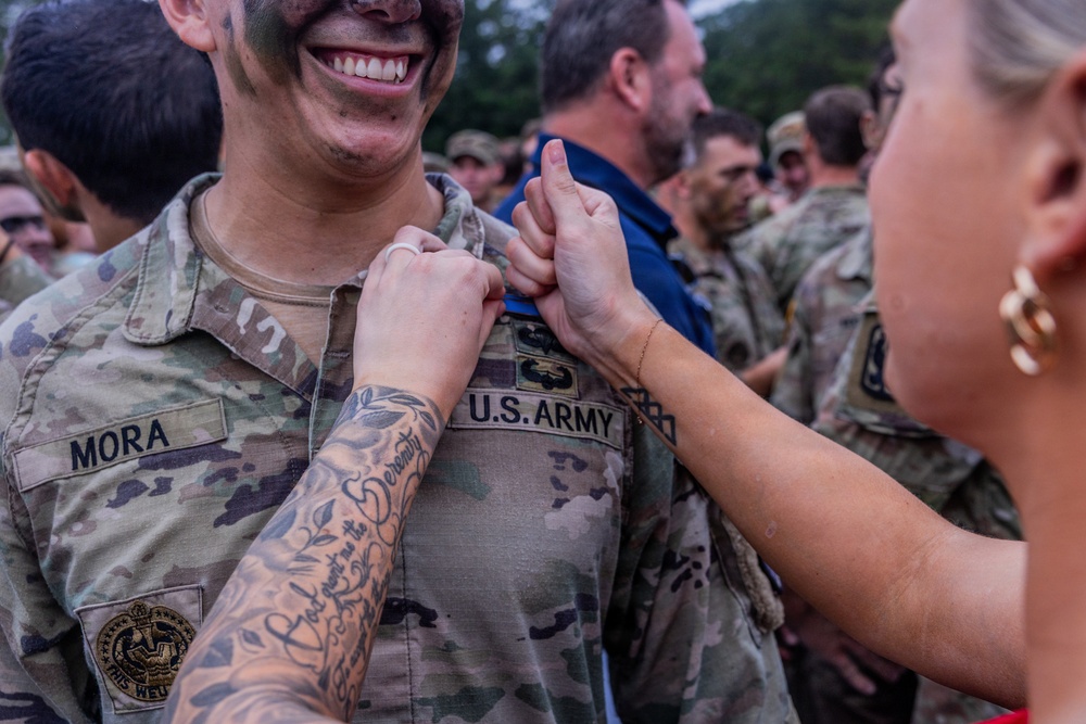 A U.S. Army soldier named Mora, wearing camouflage uniform with face paint, is smiling broadly while a woman pins something onto his uniform collar area. The scene appears to be a military pinning ceremony, likely marking a promotion or graduation. Multiple soldiers and civilians are visible in the background in a similar setting.