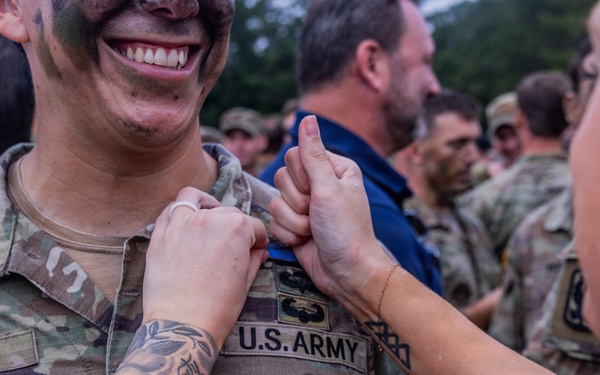 A U.S. Army soldier named Mora, wearing camouflage uniform with face paint, is smiling broadly while a woman pins something onto his uniform collar area. The scene appears to be a military pinning ceremony, likely marking a promotion or graduation. Multiple soldiers and civilians are visible in the background in a similar setting.