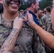 A U.S. Army soldier named Mora, wearing camouflage uniform with face paint, is smiling broadly while a woman pins something onto his uniform collar area. The scene appears to be a military pinning ceremony, likely marking a promotion or graduation. Multiple soldiers and civilians are visible in the background in a similar setting.