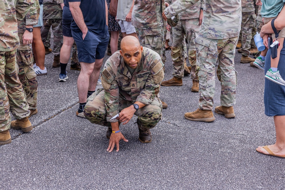 A US Army soldier in OCP (Operational Camouflage Pattern) uniform is crouched down in a starting position on a paved surface, appearing to be preparing for a physical fitness activity or race. The soldier's name tape reads 'ALLEN' and displays US ARMY insignia with sergeant chevrons visible. The scene is surrounded by a mix of other uniformed military personnel and civilians in casual clothing.