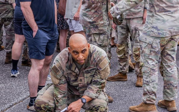 A US Army soldier in OCP (Operational Camouflage Pattern) uniform is crouched down in a starting position on a paved surface, appearing to be preparing for a physical fitness activity or race. The soldier's name tape reads 'ALLEN' and displays US ARMY insignia with sergeant chevrons visible. The scene is surrounded by a mix of other uniformed military personnel and civilians in casual clothing.
