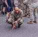 A US Army soldier in OCP (Operational Camouflage Pattern) uniform is crouched down in a starting position on a paved surface, appearing to be preparing for a physical fitness activity or race. The soldier's name tape reads 'ALLEN' and displays US ARMY insignia with sergeant chevrons visible. The scene is surrounded by a mix of other uniformed military personnel and civilians in casual clothing.