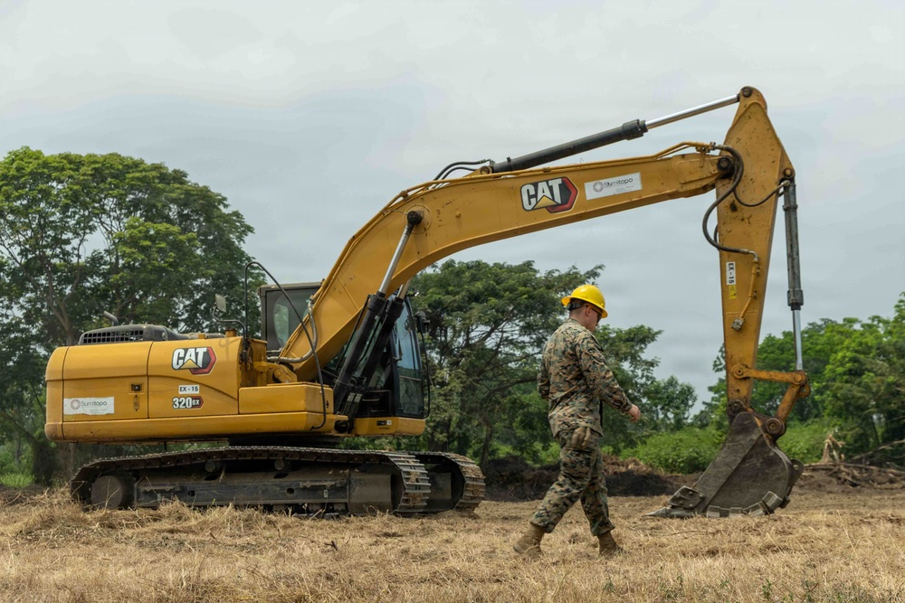 A military service member wearing camouflage uniform and a yellow hard hat walks near a large yellow Caterpillar (CAT) 320GX tracked excavator on a cleared dirt field. The setting appears to be an outdoor construction or engineering operation, likely a military civil engineering or humanitarian assistance project. Trees and overcast skies form the background.