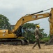 A military service member wearing camouflage uniform and a yellow hard hat walks near a large yellow Caterpillar (CAT) 320GX tracked excavator on a cleared dirt field. The setting appears to be an outdoor construction or engineering operation, likely a military civil engineering or humanitarian assistance project. Trees and overcast skies form the background.