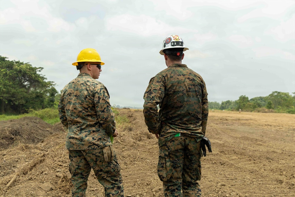 Two individuals wearing U.S. Marine Corps MARPAT digital camouflage uniforms and hard hats are standing on a cleared, earthen construction or engineering site. One wears a yellow hard hat and sunglasses, while the other wears a white hard hat with visible insignia. They appear to be overseeing or surveying a land-clearing or construction operation in a tropical or subtropical environment.